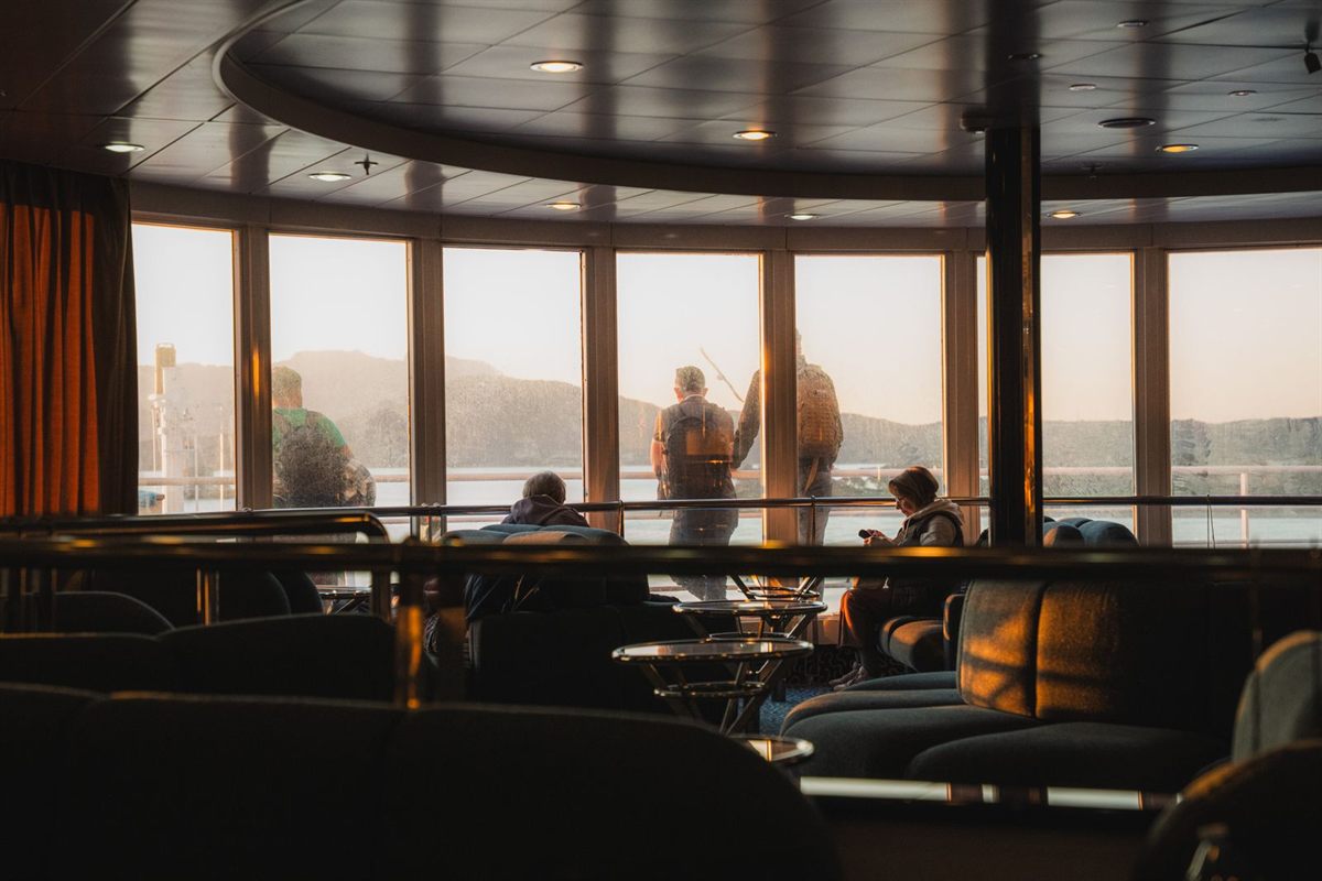 Passengers looking out of the wide windows of a ship lounge at sunset